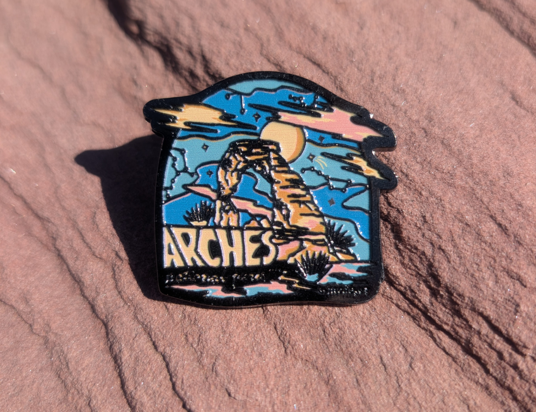 Die cut Arches enamel pin with Delicate Arch under a blue and pink starry sky, lying on red sandstone in Moab, Utah.
