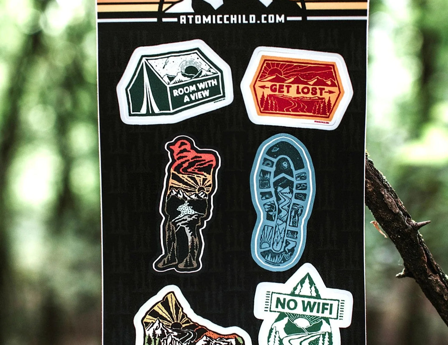Atomicchild Hiking Mini Sticker Sheet held up against a forest background