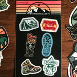 Atomicchild Hiking Mini Sticker Sheet with tent, hiking boot, No WiFi and Get Lost stickers on a wooden table