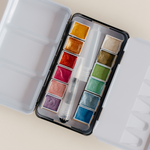 Bailey Rae Studio 12 Color Travel Metallic Watercolor Paint Palette, open tin showing twelve shimmering metallic watercolor pans and a brush
