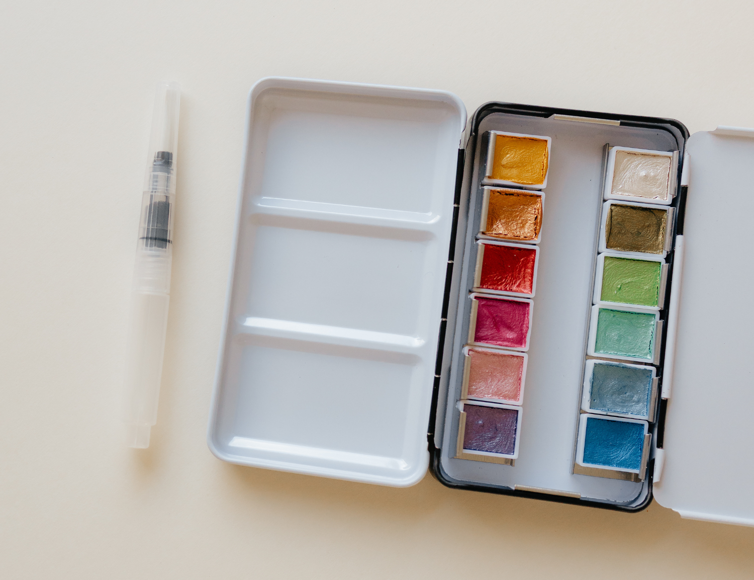 Bailey Rae Studio 12 Color Travel Metallic Watercolor Paint Palette, open silver tin with metallic pans and a water brush pen on a cream background