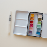 Bailey Rae Studio 12 Color Travel Metallic Watercolor Paint Palette, open silver tin with metallic pans and a water brush pen on a cream background