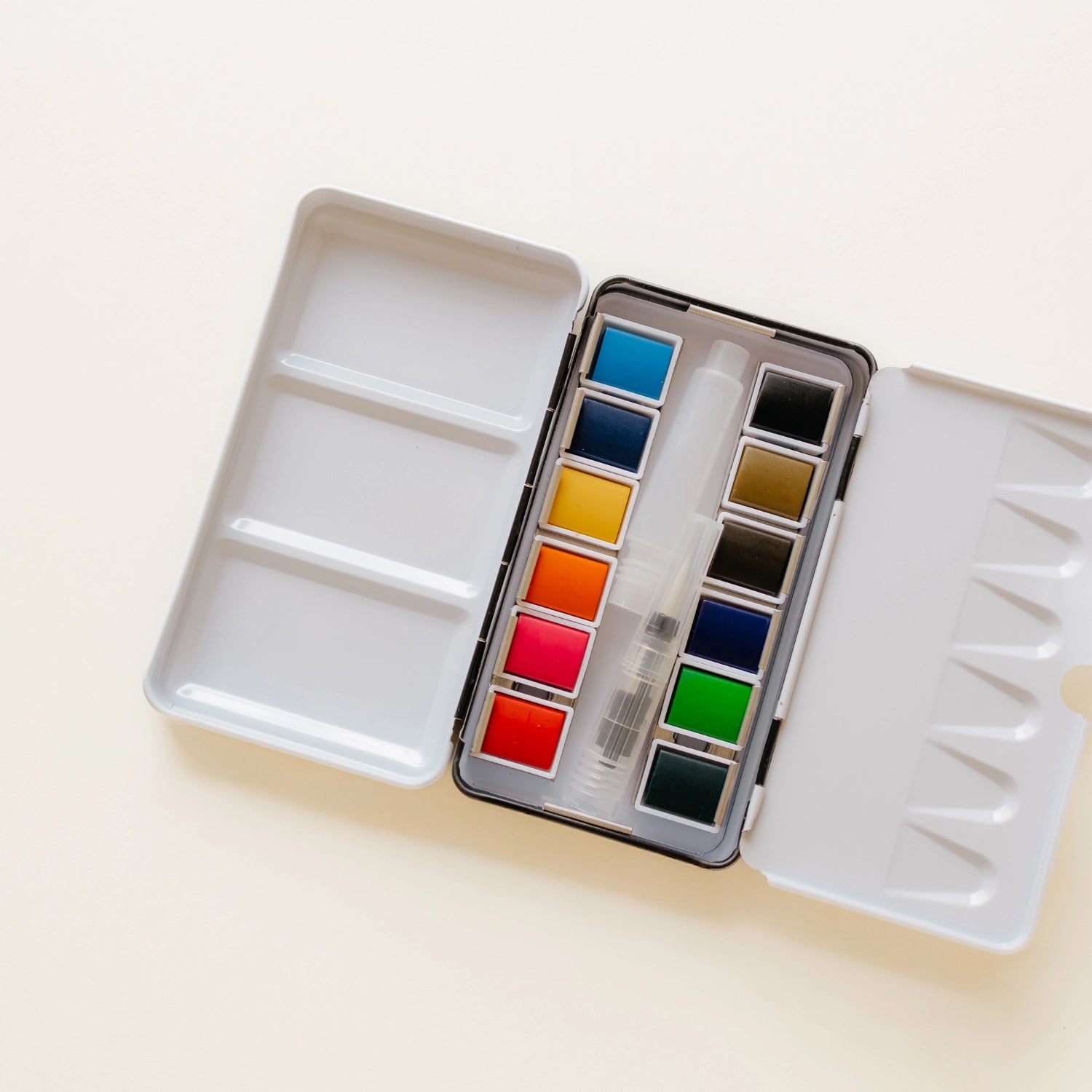 Bailey Rae Studio 12 Color Mini-Travel Paint Palette, open silver tin with twelve watercolor pans in blues, greens, reds, and yellows on a cream background
