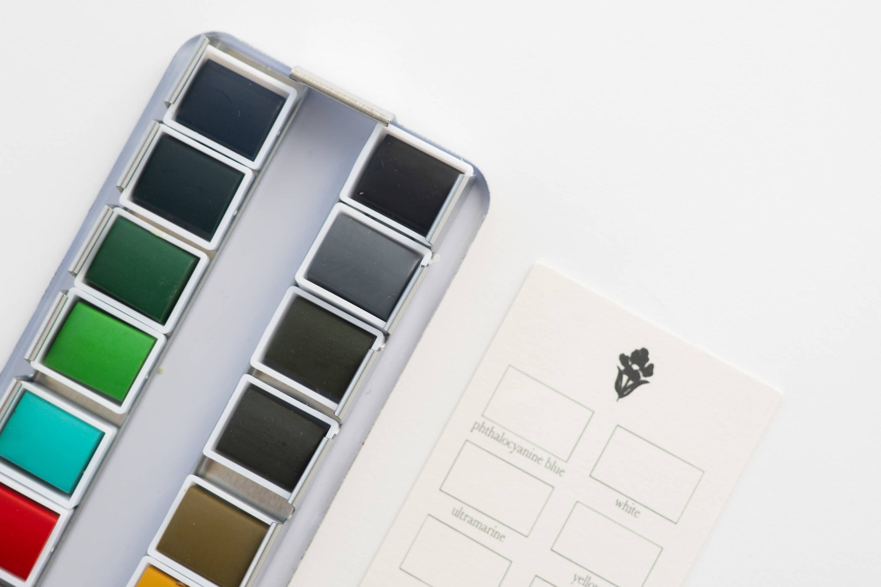 Bailey Rae Studio 24 Color Watercolor Paint Palette, close-up of green, teal, brown and grey pans beside the swatch chart card