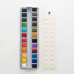 Bailey Rae Studio 24 Color Watercolor Paint Palette, open tin of colorful pans next to a blank color swatch chart card, on white