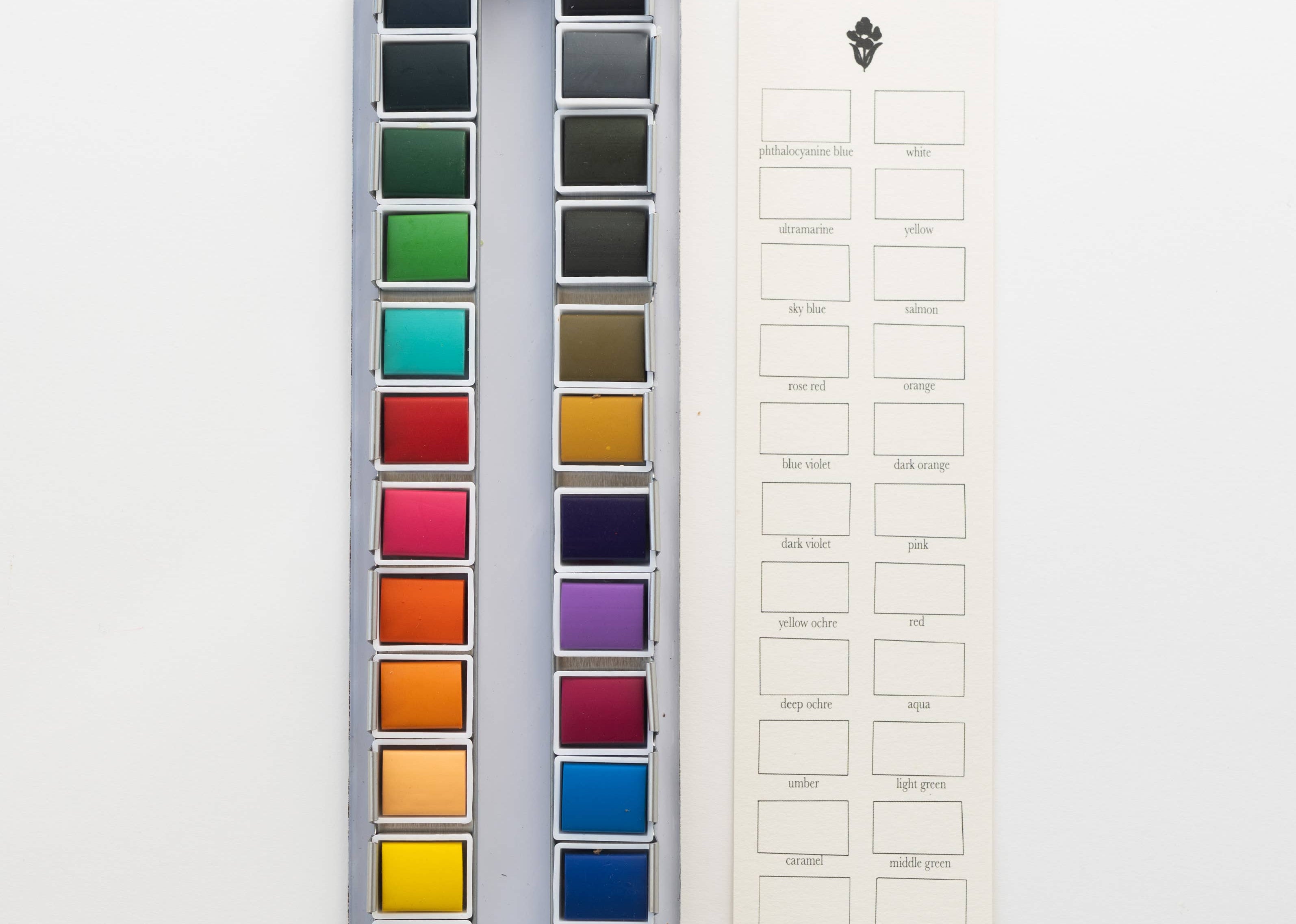 Bailey Rae Studio 24 Color Watercolor Paint Palette, open tin of colorful pans next to a blank color swatch chart card, on white