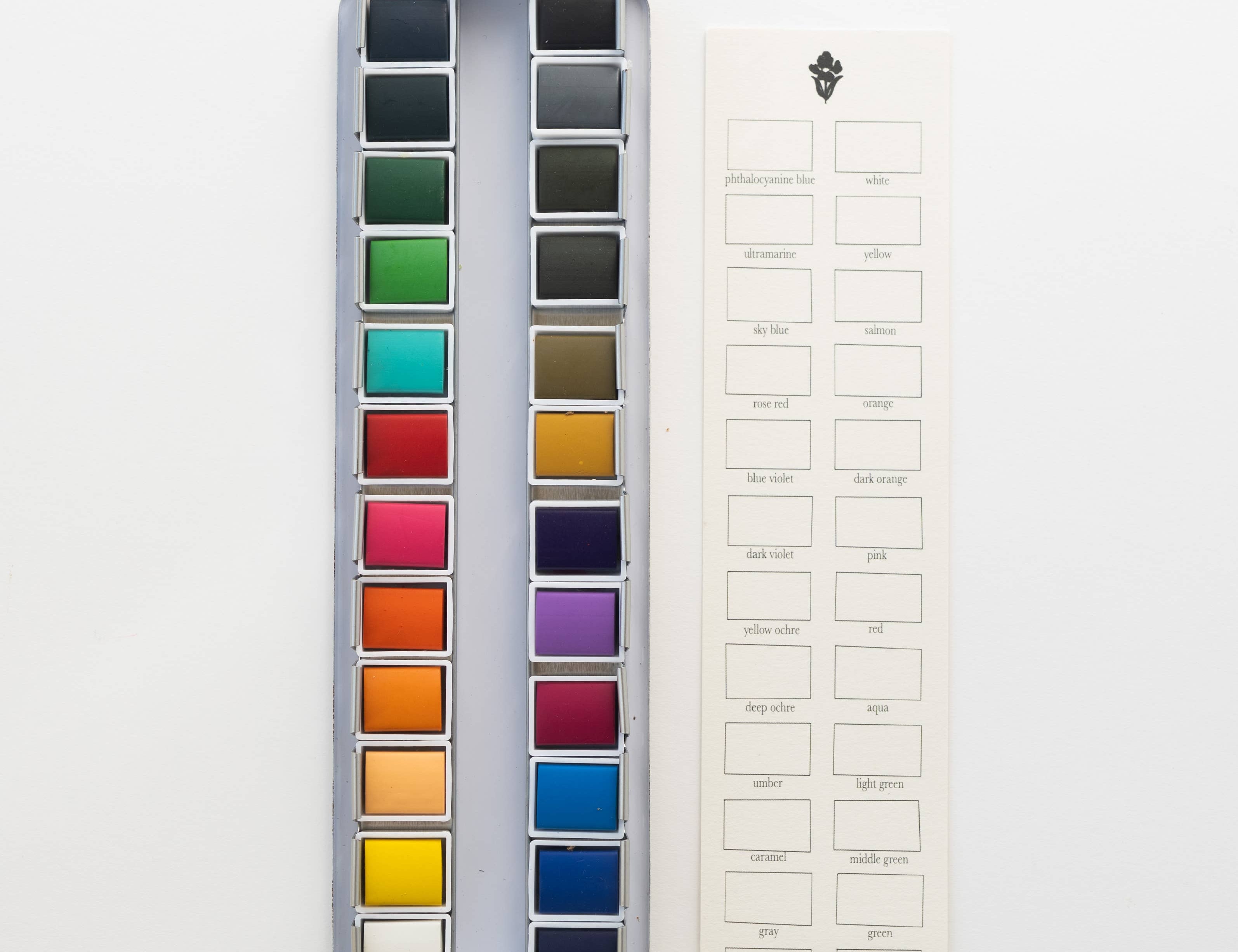 Bailey Rae Studio 24 Color Watercolor Paint Palette, open tin of colorful pans next to a blank color swatch chart card, on white