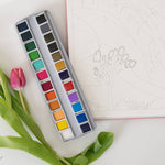 Bailey Rae Studio 24 Color Watercolor Paint Palette, open tin on a table with a pink tulip and a watercolor outline page