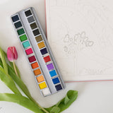 Bailey Rae Studio 24 Color Watercolor Paint Palette, open tin on a table with a pink tulip and a watercolor outline page