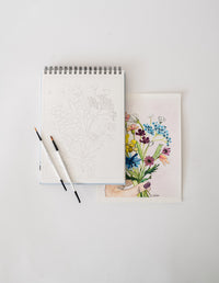 Bailey Rae Studio Floral Watercoloring Book open to an outline page with brushes and a finished floral painting