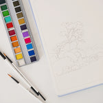 Bailey Rae Studio Landscape Watercoloring Book open to a tree outline with a paint palette and brushes