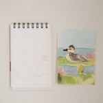 Bailey Rae Studio Mini A Little Bit of Spring Watercoloring Book, bird outline page beside a painted shorebird on a pond