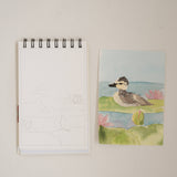 Bailey Rae Studio Mini A Little Bit of Spring Watercoloring Book, bird outline page beside a painted shorebird on a pond
