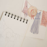Bailey Rae Studio Mini A Little Bit of Spring Watercoloring Book, clothesline outline page beside a painted line of dresses and a straw hat
