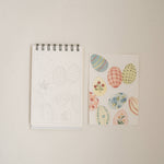 Bailey Rae Studio Mini A Little Bit of Spring Watercoloring Book, Easter egg outline page beside painted pastel patterned eggs