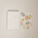 Bailey Rae Studio Mini A Little Bit of Spring Watercoloring Book, Easter egg outline page beside painted pastel patterned eggs