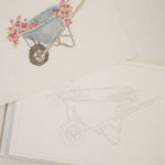 Bailey Rae Studio Mini A Little Bit of Spring Watercoloring Book, wheelbarrow outline page beside a painted wheelbarrow of pink flowers