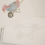 Bailey Rae Studio Mini A Little Bit of Spring Watercoloring Book, wheelbarrow outline page beside a painted wheelbarrow of pink flowers