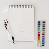 Bailey Rae Studio National Parks Watercoloring Book open to a canyon outline with a paint palette and brushes