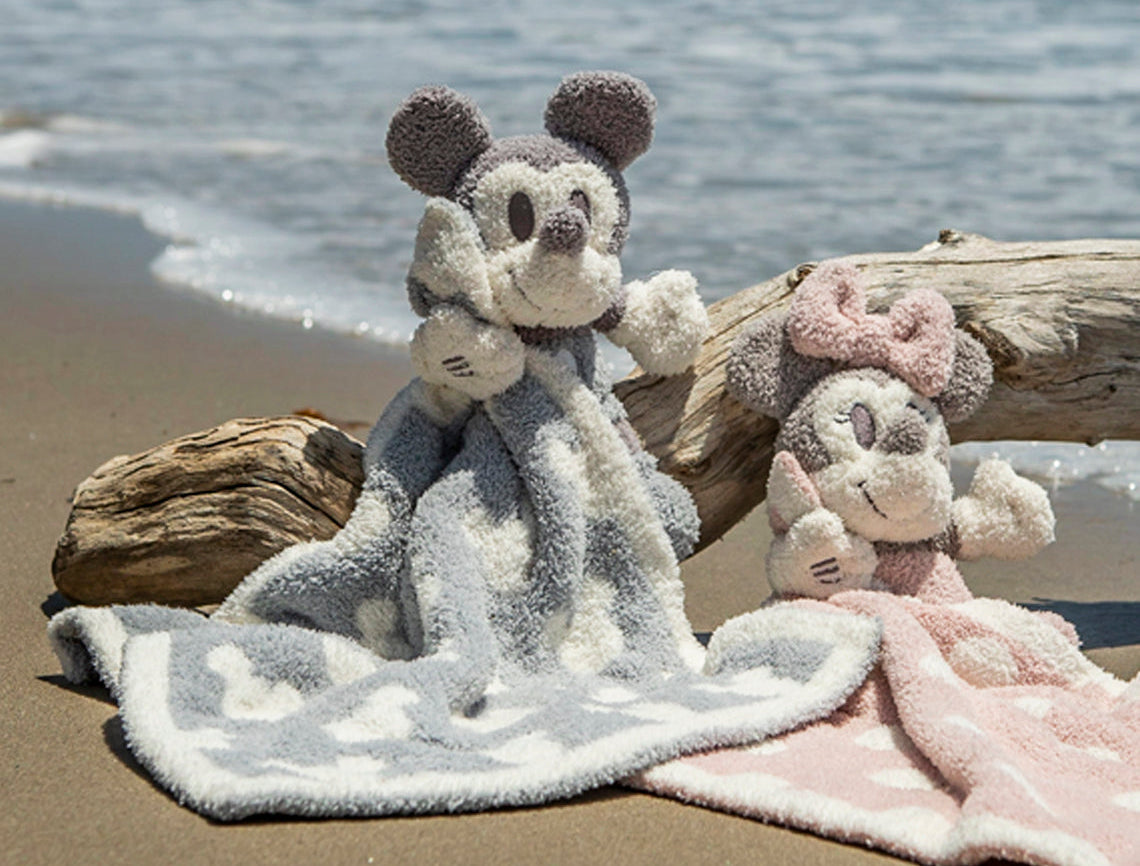 Barefoot Dreams Disney Blanket Buddies in Vintage Mickey and Vintage Minnie on a beach beside driftwood