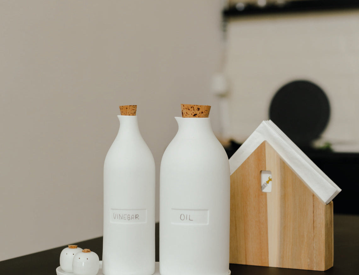 Blomus Bon Appetit Porcelain Cruet Bottles on a dark counter beside a small wooden house-shaped board