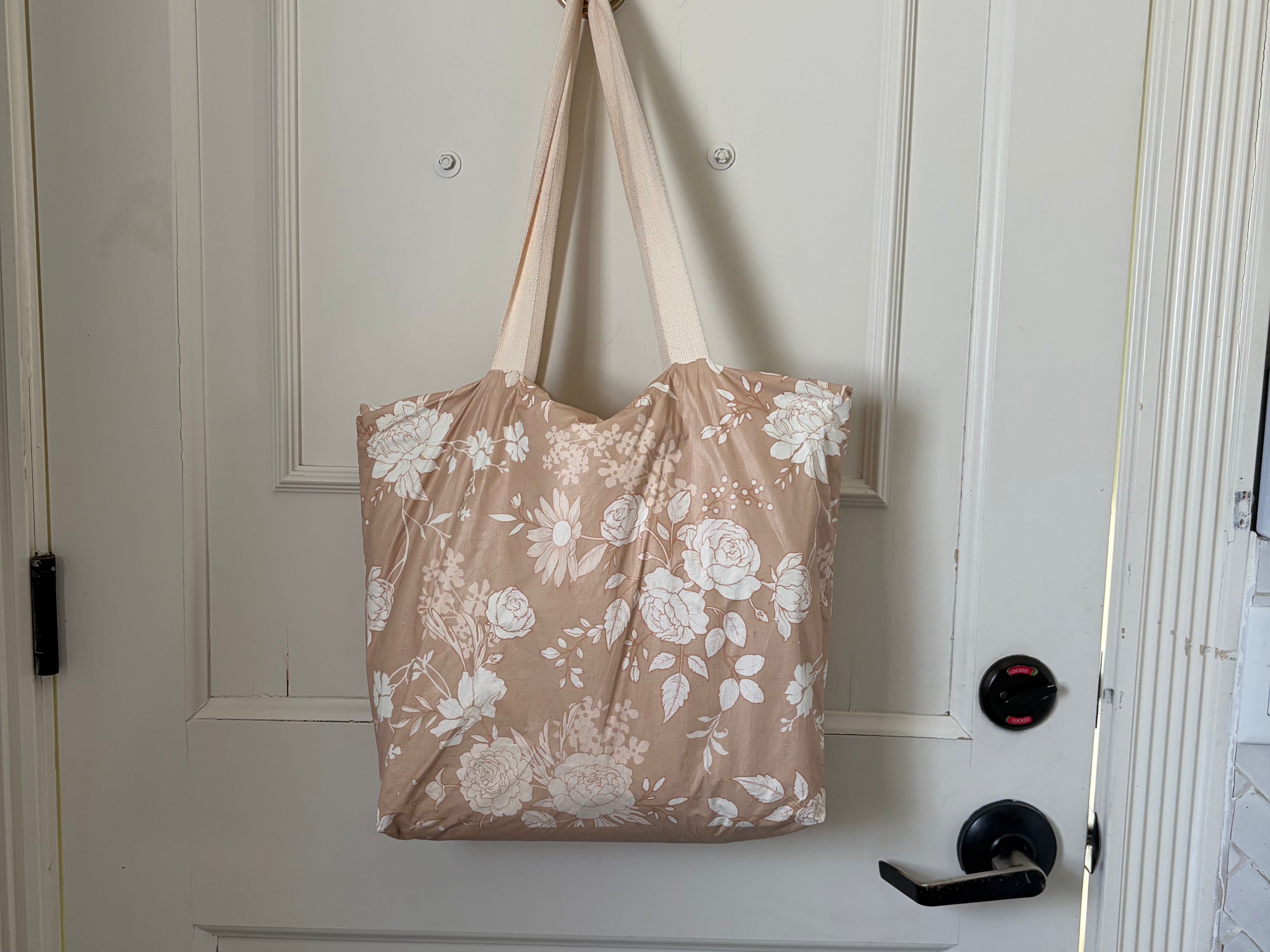 The Canyon Rose tote hanging from a hook on a white panelled door, warm clay rose printed with white roses and daisies.