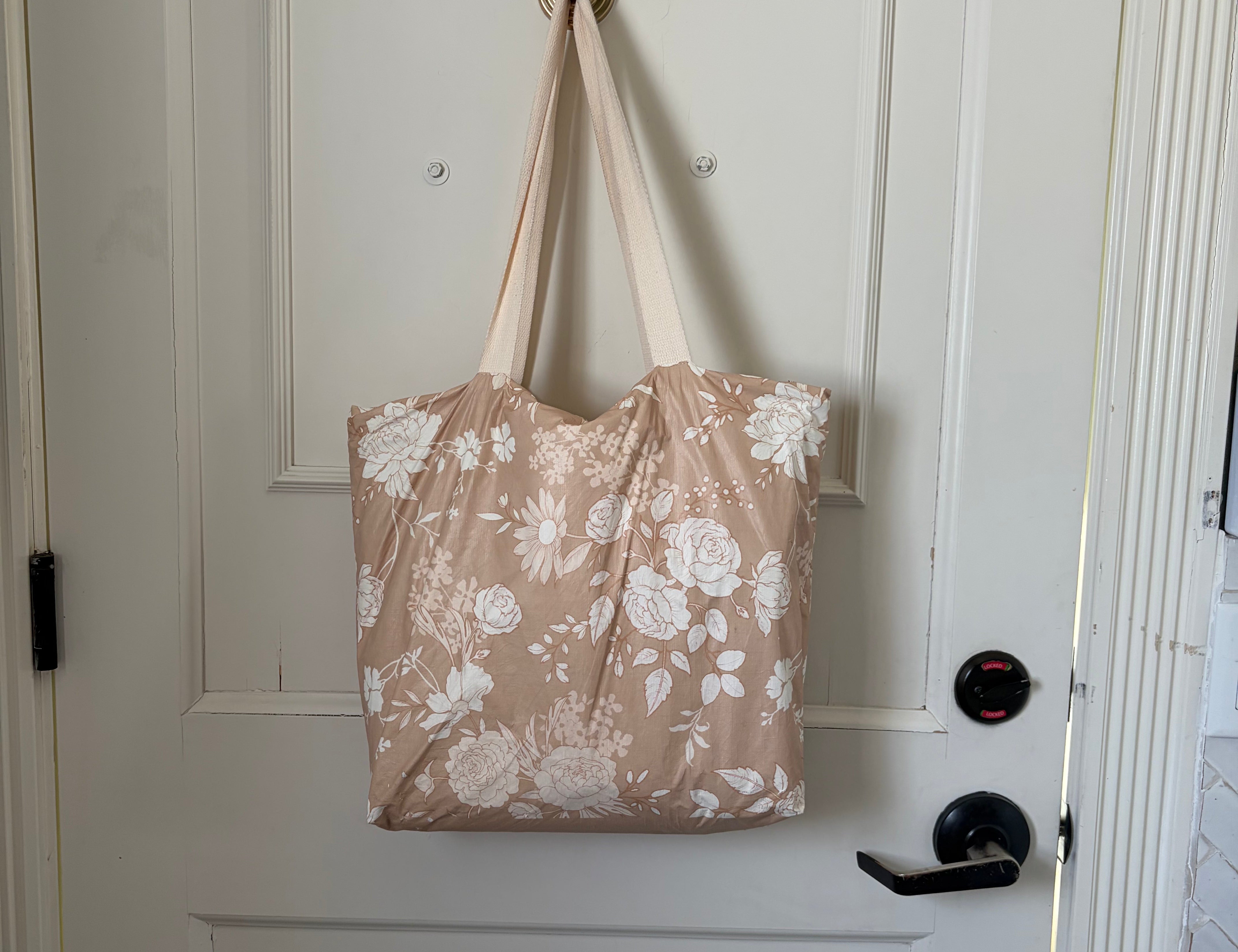 The Canyon Rose tote hanging from a hook on a white panelled door, warm clay rose printed with white roses and daisies.