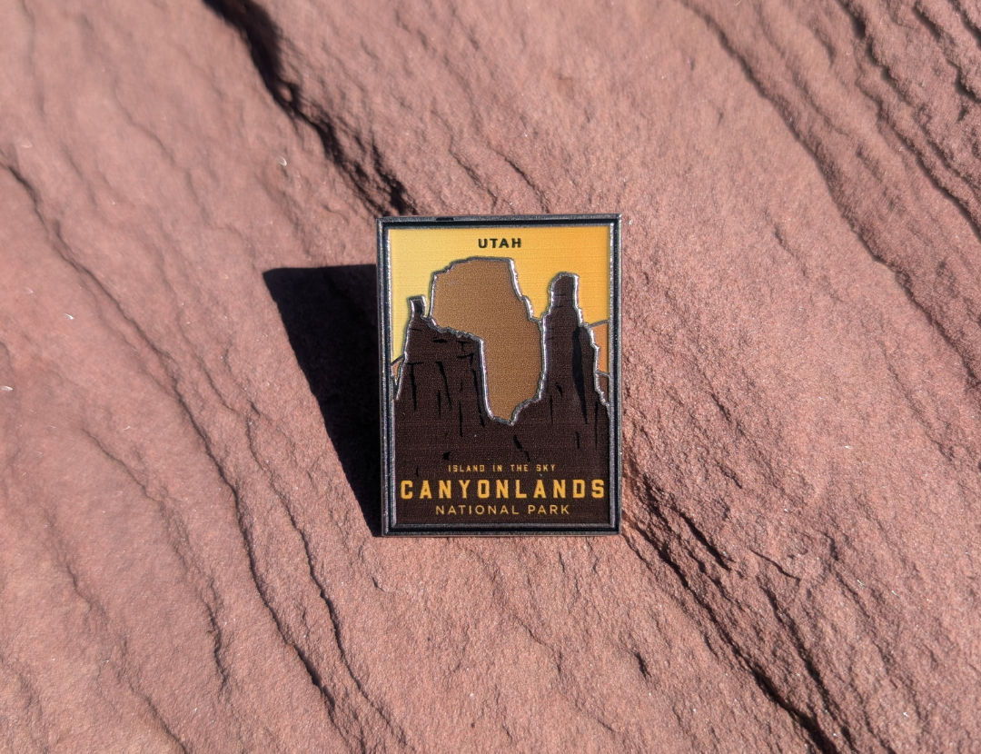 Canyonlands National Park Island in the Sky enamel pin with dark buttes against a golden sky, lying on red sandstone in Moab, Utah.