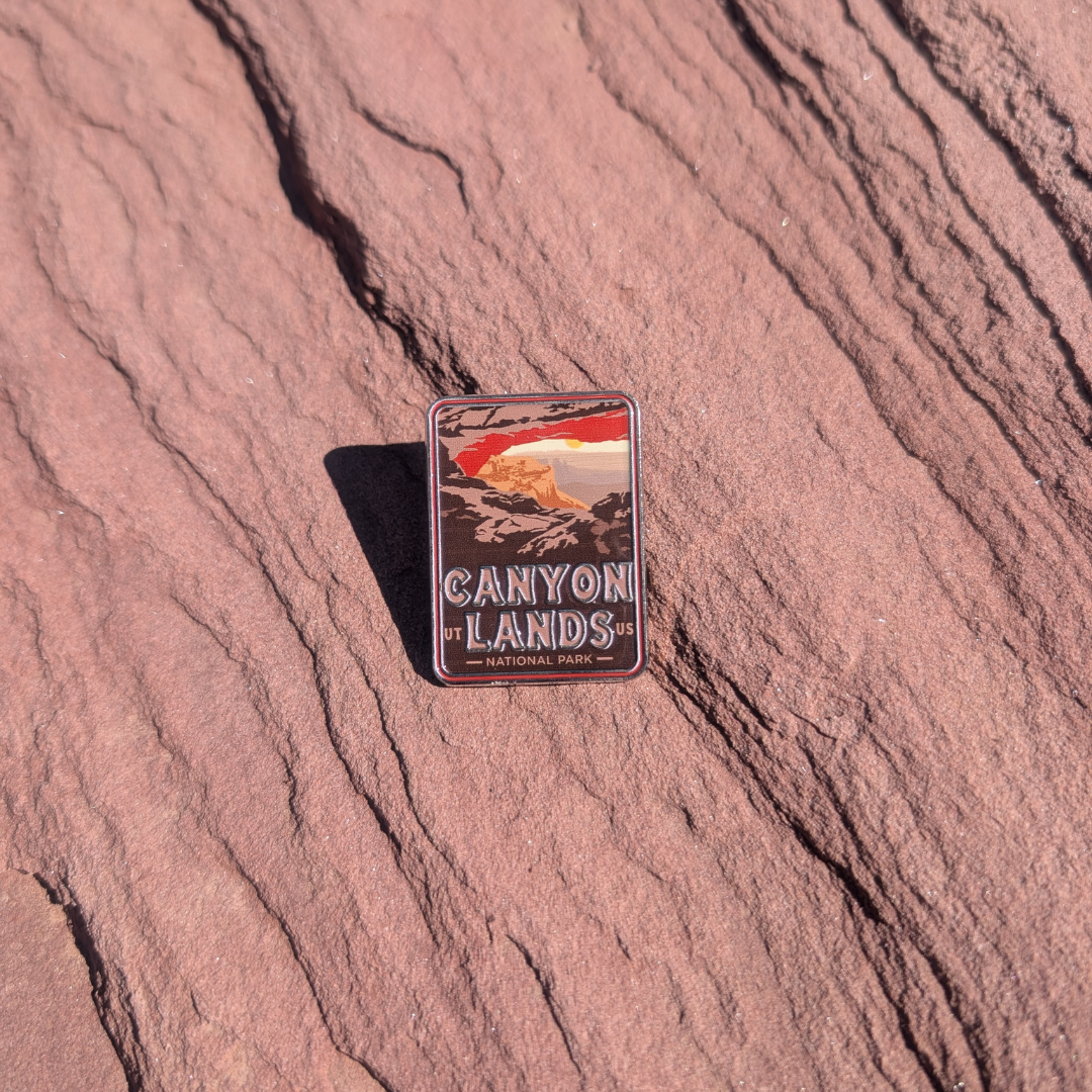 Canyonlands National Park enamel pin framing a sunrise through Mesa Arch, lying on red sandstone in Moab, Utah.