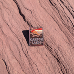 Canyonlands National Park enamel pin framing a sunrise through Mesa Arch, lying on red sandstone in Moab, Utah.