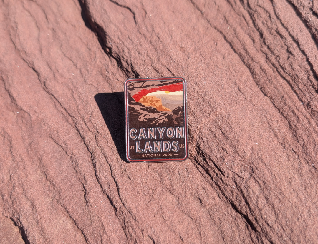 Canyonlands National Park enamel pin framing a sunrise through Mesa Arch, lying on red sandstone in Moab, Utah.