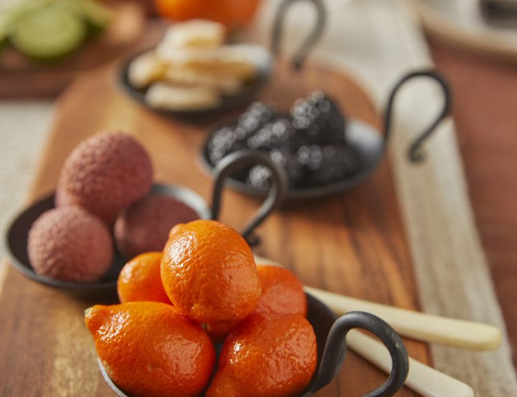 Multiple Handmade Forged Cast Iron Serving Dishes with black matte finish and thin curving handle, holding variety of exotic product on cutting board. 