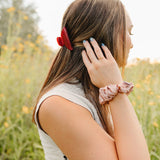 The Garnet Classic claw clip worn with a blush silk scrunchie on the wrist, in a field at golden hour.