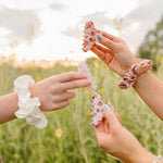 Three hands holding up floral and pearl Classic claw clips and silk scrunchies against the evening sky.