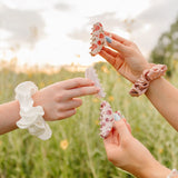 Three hands holding up floral and pearl Classic claw clips and silk scrunchies against the evening sky.