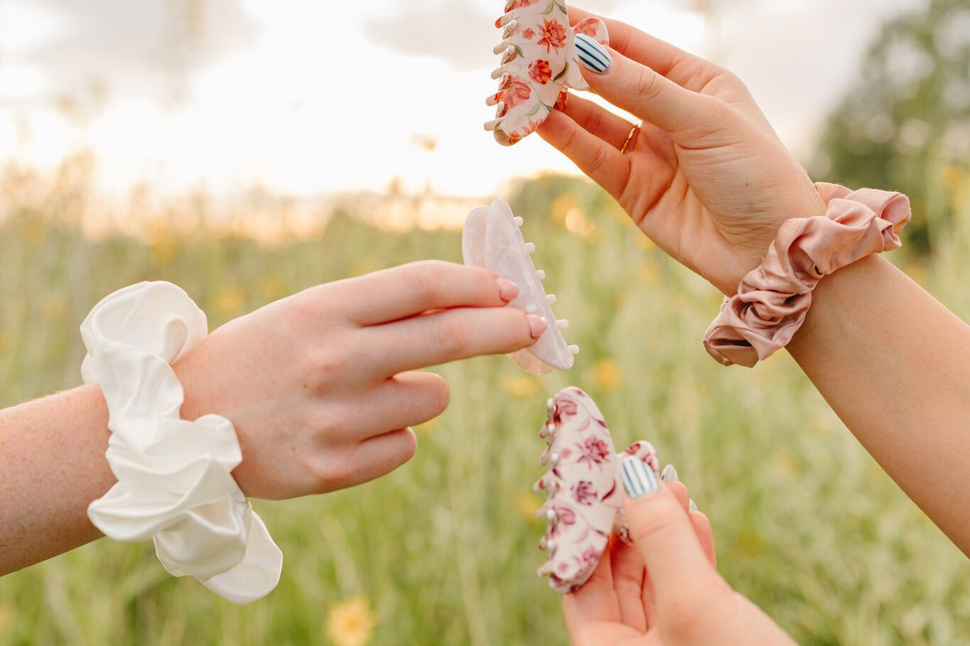 Three hands holding up floral and pearl Classic claw clips and silk scrunchies against the evening sky.