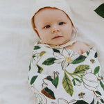 Baby swaddled in the Clementine Kids Magnolia Swaddle Blanket lying on a white bed
