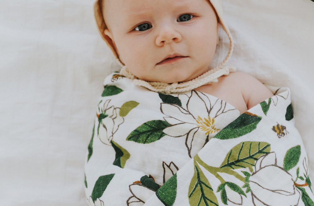 Baby swaddled in the Clementine Kids Magnolia Swaddle Blanket lying on a white bed