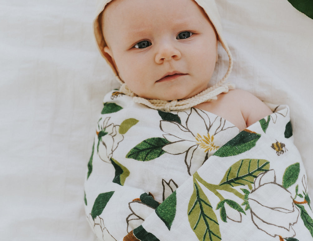 Baby swaddled in the Clementine Kids Magnolia Swaddle Blanket lying on a white bed