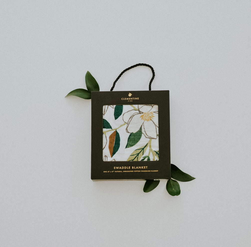 Clementine Kids Magnolia Swaddle Blanket folded in its black hang-tag packaging with a magnolia sprig