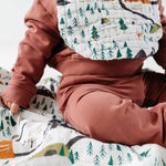 Baby in a rust outfit wearing the Clementine Kids National Park Bib, sitting on a matching blanket