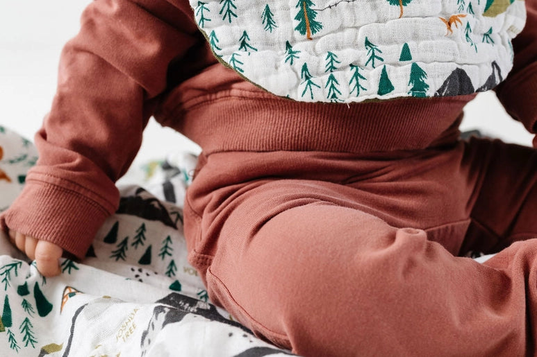 Baby in a rust outfit wearing the Clementine Kids National Park Bib, sitting on a matching blanket