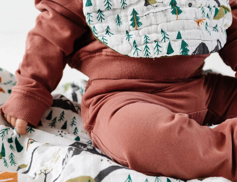 Baby in a rust outfit wearing the Clementine Kids National Park Bib, sitting on a matching blanket