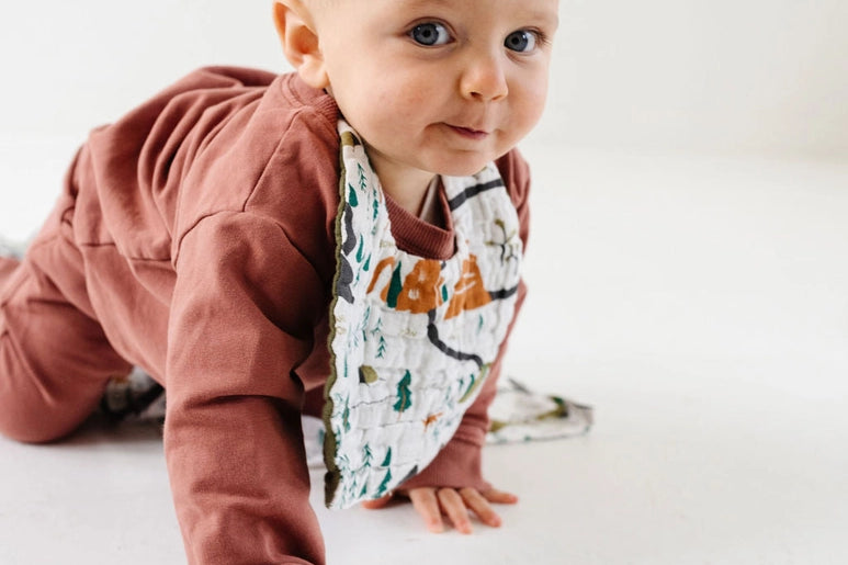 Baby in a rust outfit crawling while wearing the Clementine Kids National Park Bib