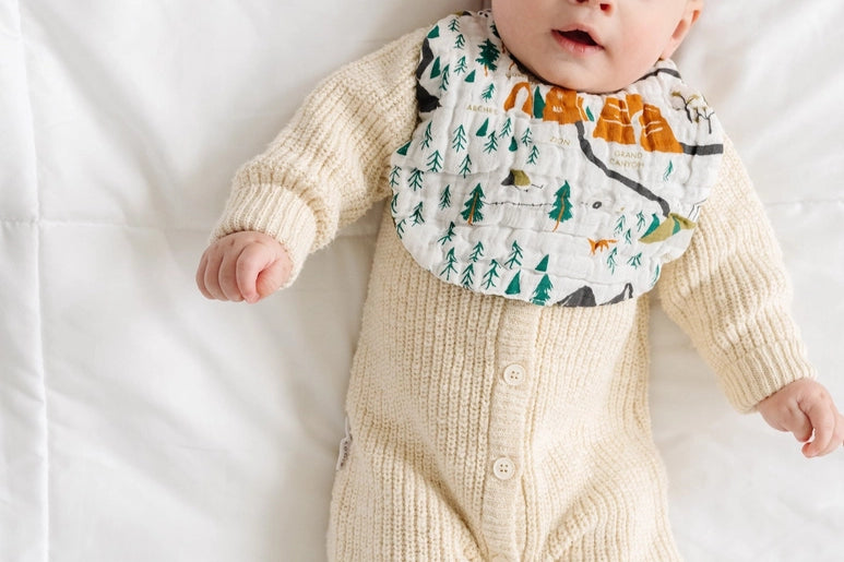 Baby in a cream knit outfit lying down wearing the Clementine Kids National Park Bib