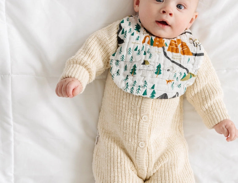 Baby in a cream knit outfit lying down wearing the Clementine Kids National Park Bib