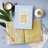 Compendium Create Your Self Guided Journal on a blue table with a candle, plants and the Self-Esteem section open