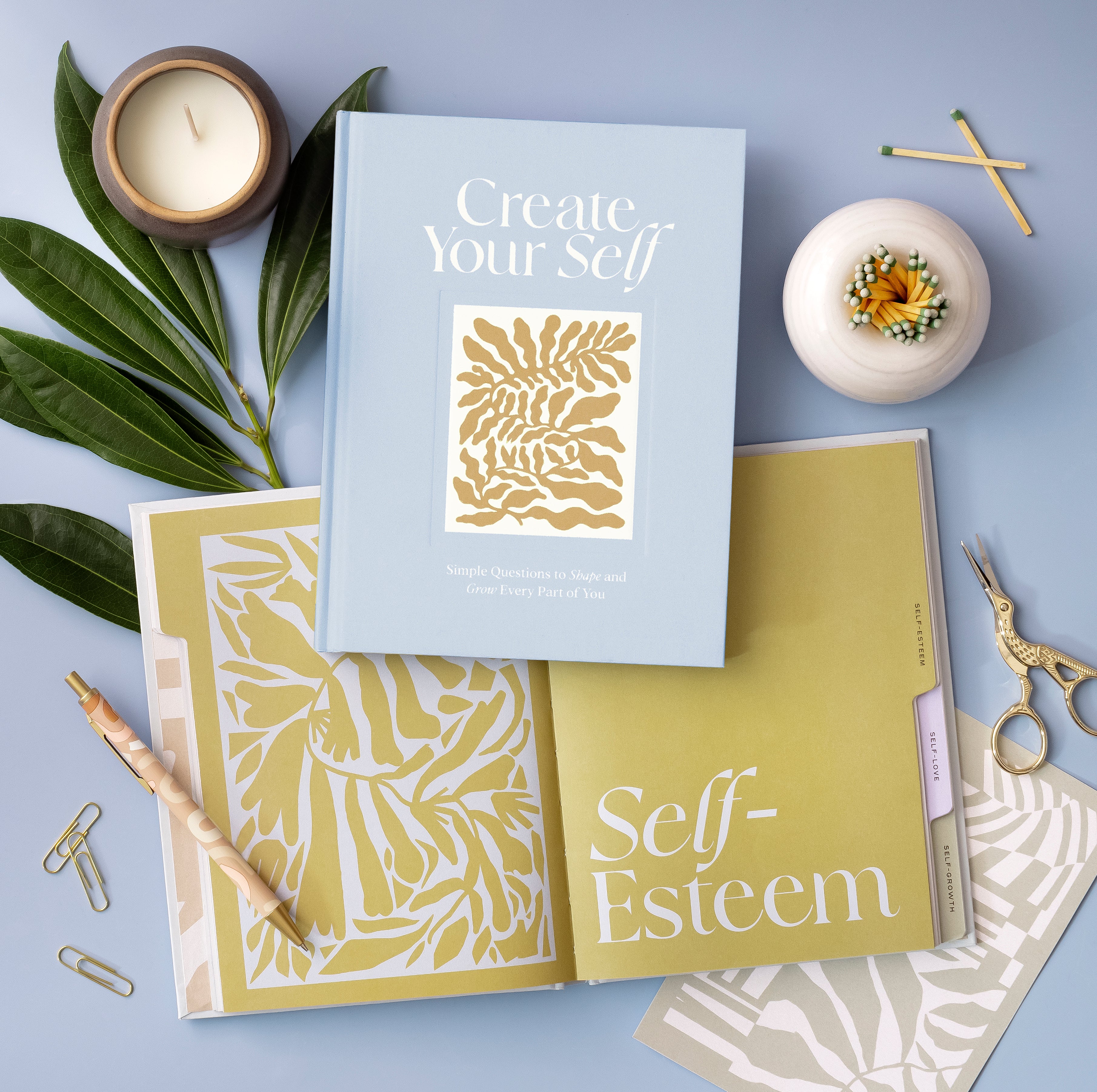 Compendium Create Your Self Guided Journal on a blue table with a candle, plants and the Self-Esteem section open