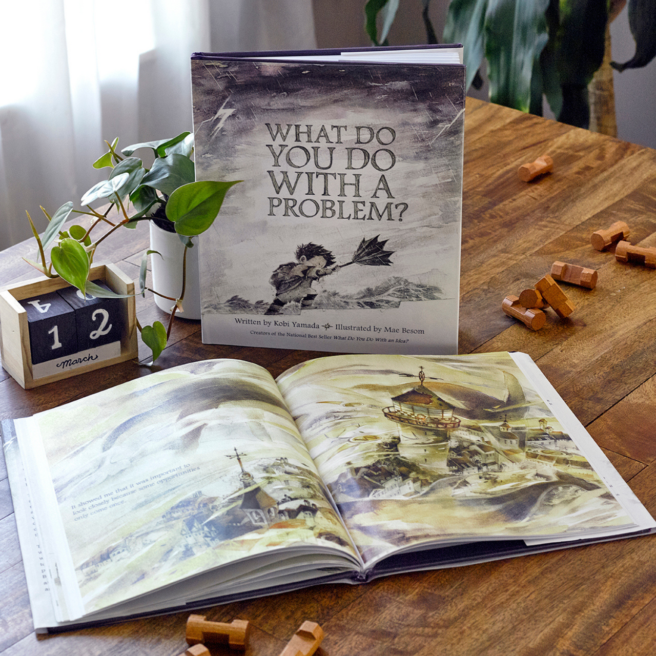 Compendium What Do You Do With a Problem book on a wooden table with an open copy and a plant