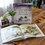 Compendium What Do You Do With a Problem book on a wooden table with an open copy and a plant